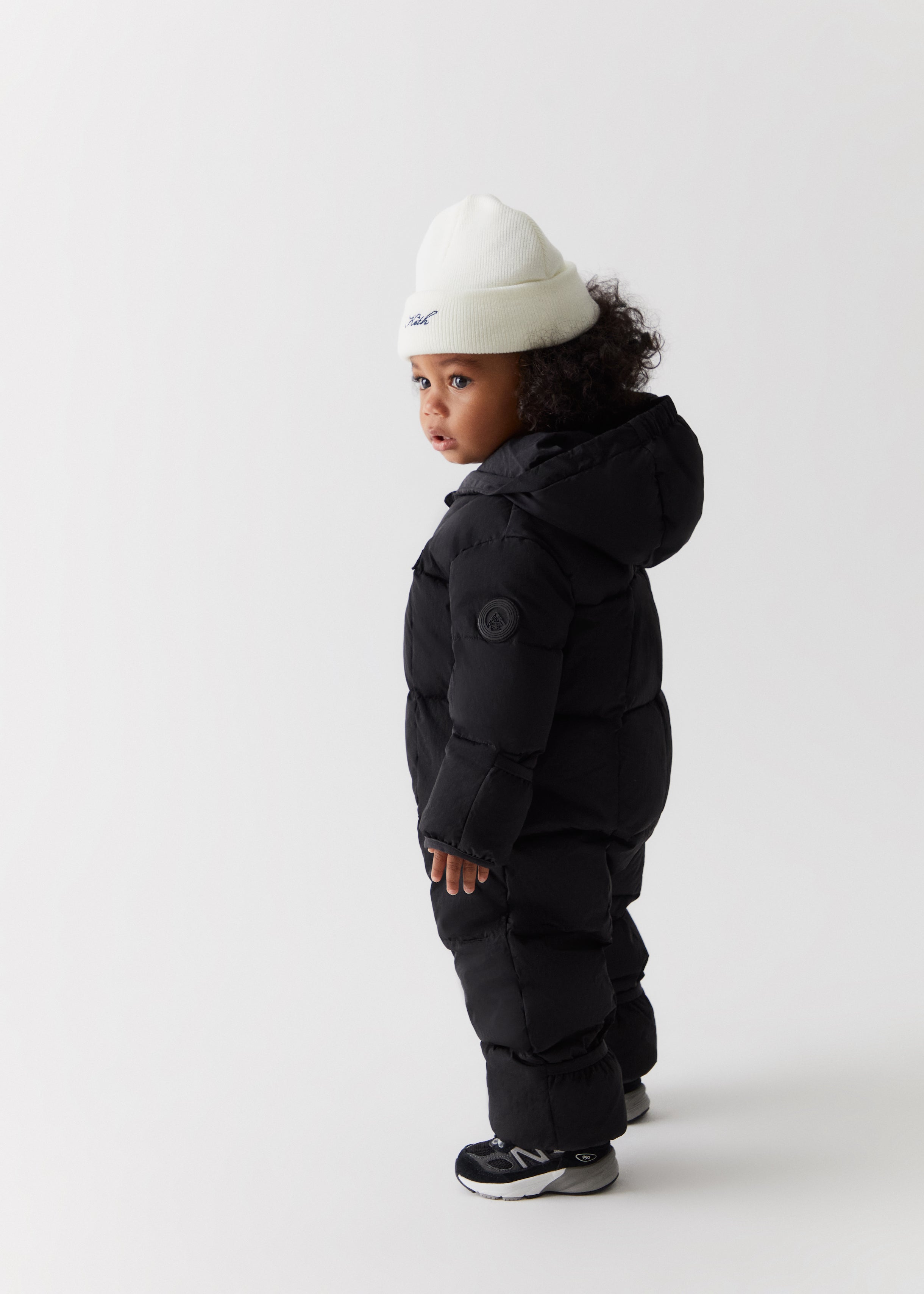 A toddler wearing a black puffer snowsuit and a Kith script knit beanie from Kith Kids Fall Classics 2023.