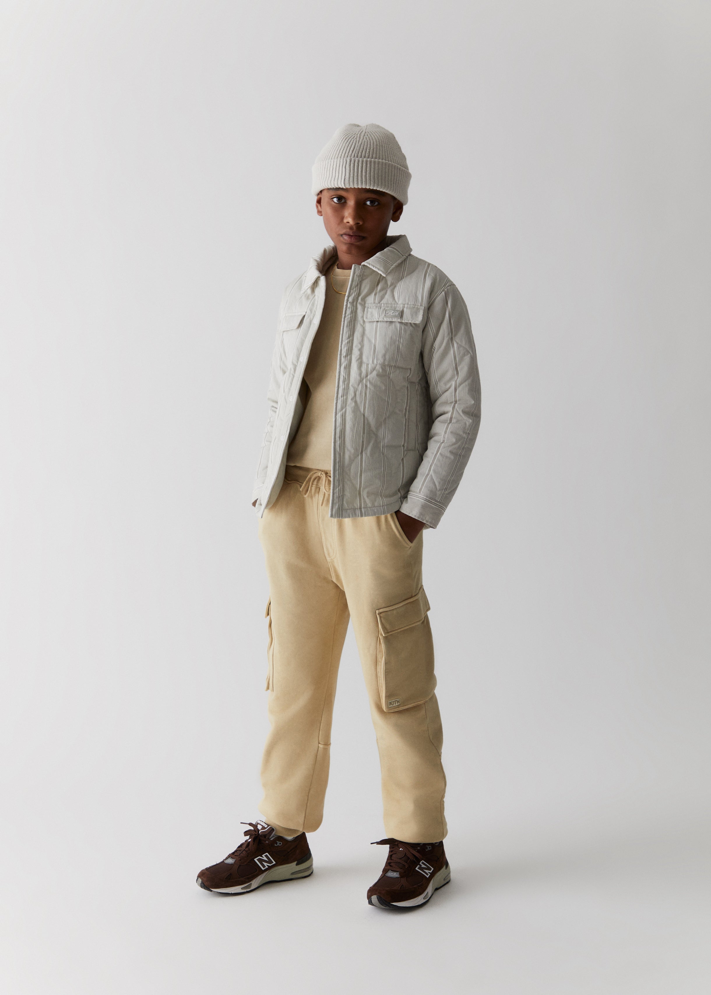 A child wearing a white knit beanie, grey quilted shirt jacket, and brown cargo pants from the Kith Kids Fall Classics 2023 collection.