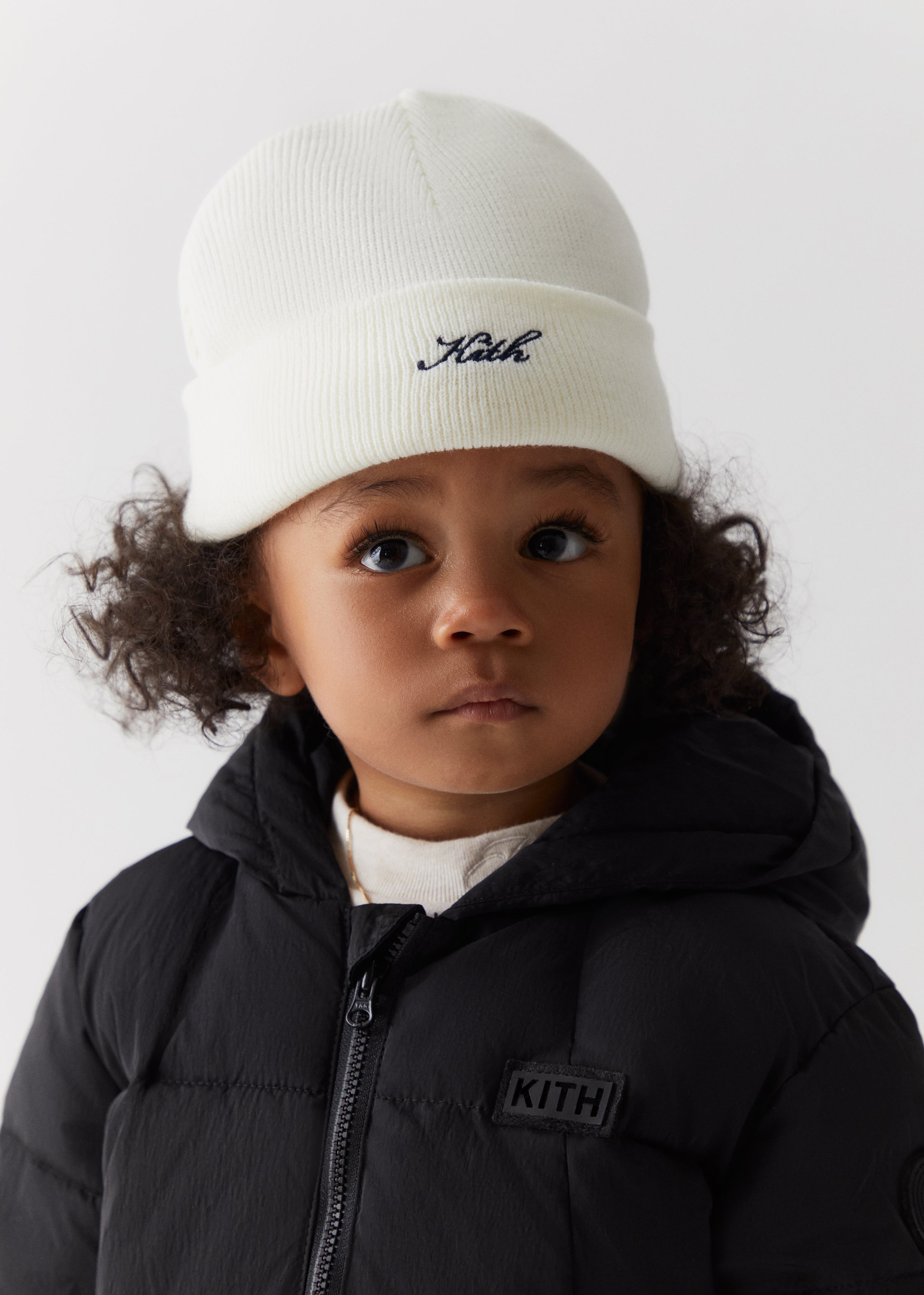 A toddler wearing a white knit beanie with blue script Kith embroidery and a black Kith puffer jacket from the Kith Kids Fall Classics 2023 collection.