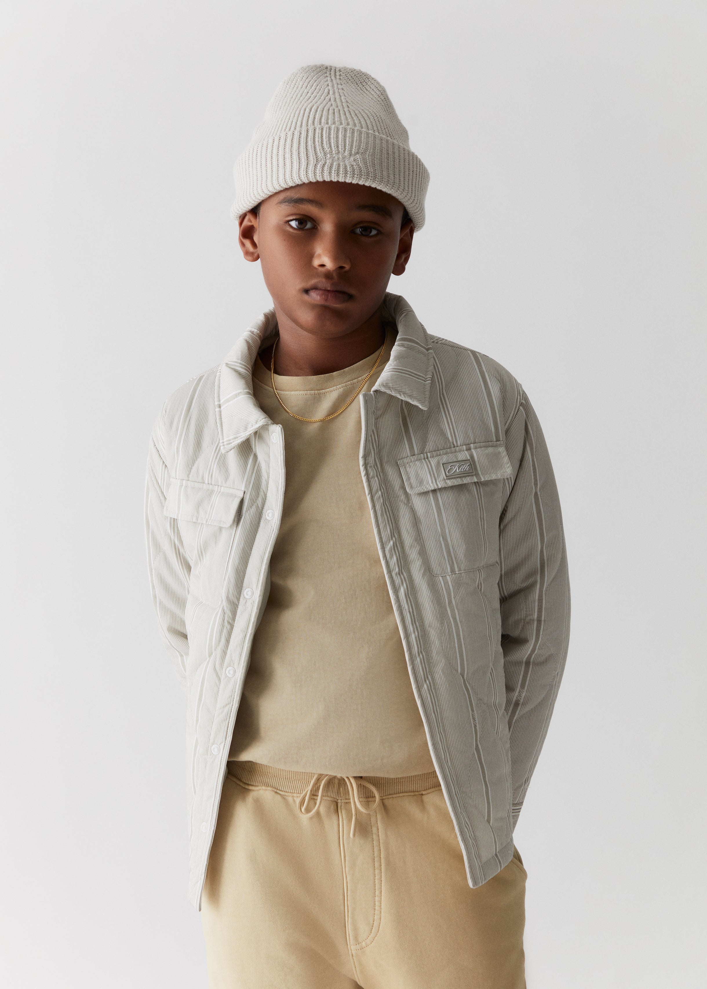A child wearing a tan knit beanie, grey striped shirt jacket, and tan cargo sweatpants from the Kith Kids Fall Classics 2023 collection.