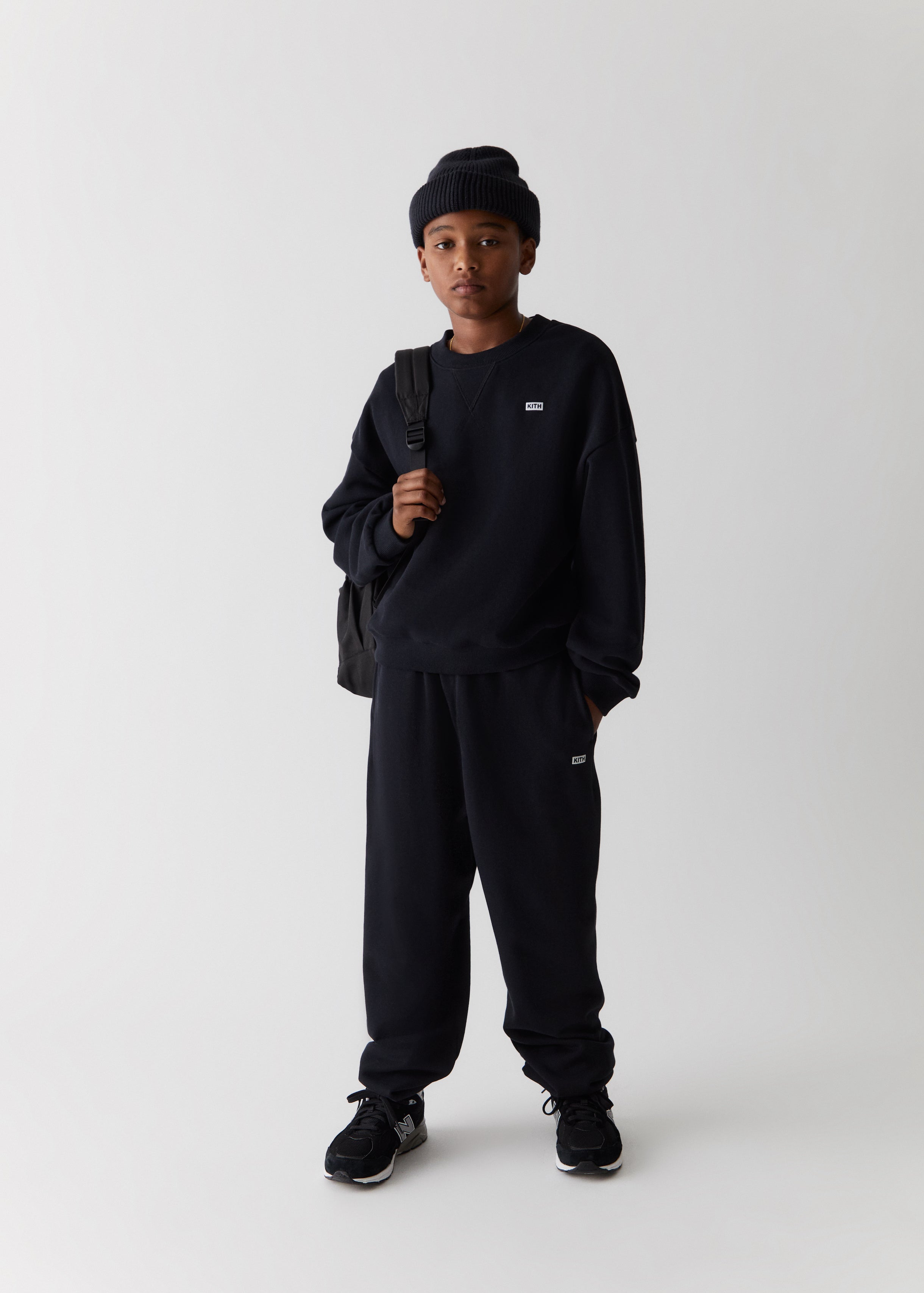 A child wearing a black knit beanie, black Kith-branded crewneck sweatshirt, black sweatpants, and black backpack from the Kith Kids Fall Classics 2023 collection.